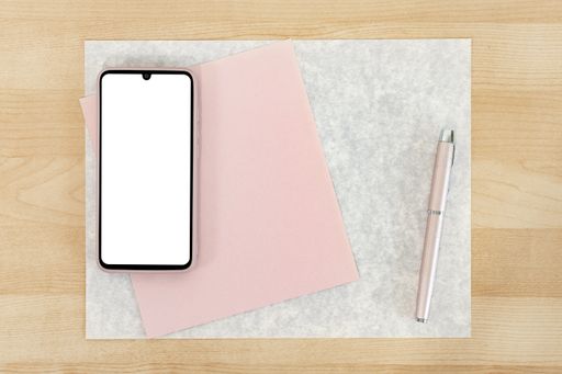 A top view shot of a wooden table with a phone over the textured, pink paper and a pen