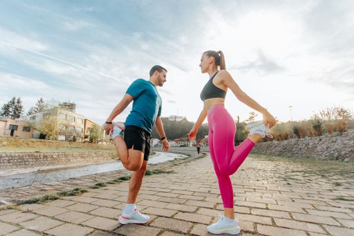 A man and a woman stretching by a riverside path under a bright sky, wearing athletic attire.