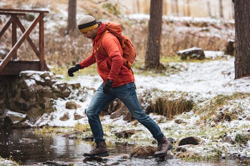 A man in his 30s, wearing winter clothing, crosses a snowy stream in a forest, showcasing outdoor adventure and exploration