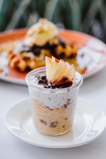 A close-up of a delicious parfait with yogurt, oats, raisins, and apple slices served in a glass jar on a white plate.