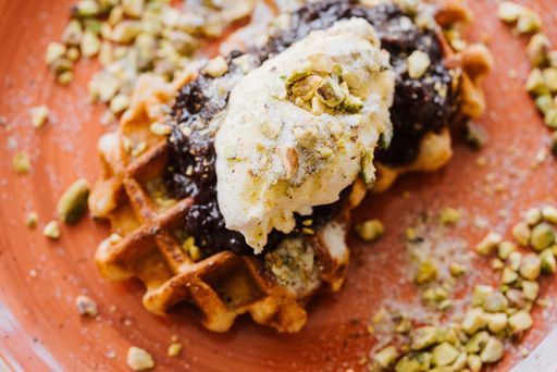Close-up of a waffle topped with cream and pistachios on a brown plate, surrounded by scattered nuts.