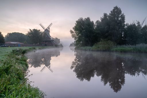Scenic view of a windmill by a tranquil river with trees reflected in the water during a misty morning.
