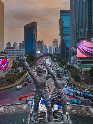 Aerial view of Jakarta's iconic Selamat Datang Monument with bustling traffic and skyscrapers under a cloudy sky.