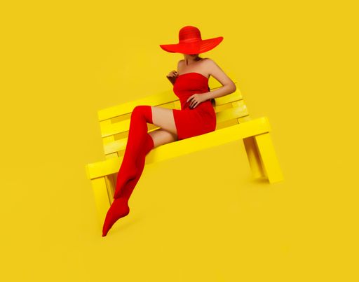 A female in a mini red dress with long red stockings and a hat sitting on a yellow bench isolated on a yellow background