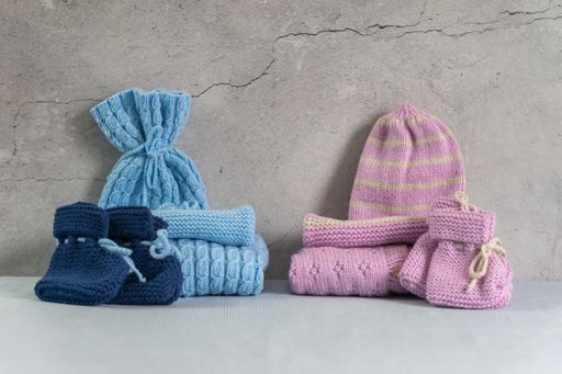 Stack of warm baby sweaters, caps, socks and pants, knitted clothes on a table. Blue and pink homemade garment for twins. Gray background, copy space.