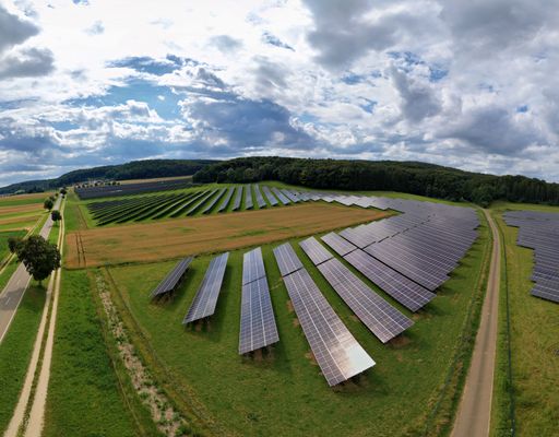 Aerial view of a large solar panel farm in a rural landscape with a cloudy sky, showcasing renewable energy technology.