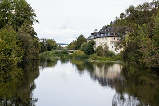 Scenic view of a tranquil river with lush greenery and a building on the riverside under a cloudy sky.