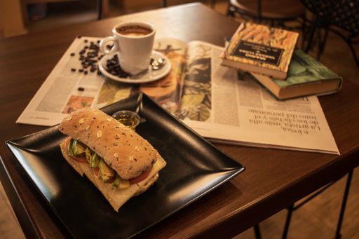 A closeup shot of a coffee cup with beans  and a sandwich near book and newspaper on the table