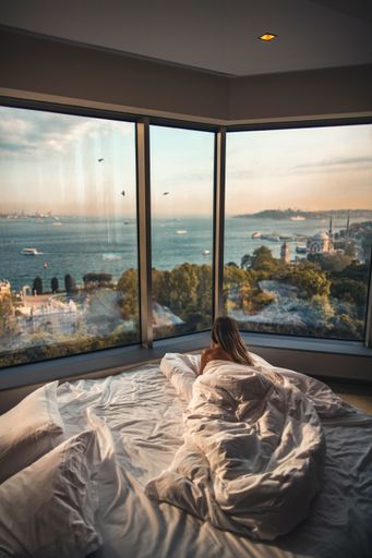 A rear view of a female lying on bed looking at the beach through the glass windows