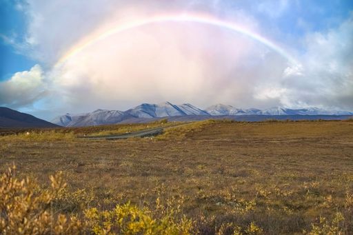 Rainbow arches over a vast landscape in Tombstone Park, Yukon, Canada. A road winds through a brown field, with mountains in the distance.