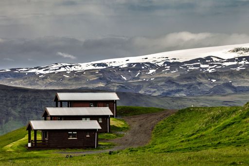 Scenic view of wooden cabins on a grassy hill with a snow-capped mountain in the background under a cloudy sky
