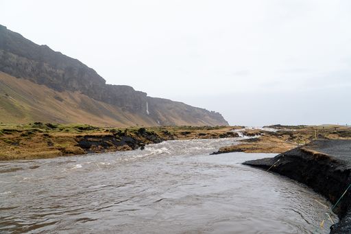 A scenic landscape of a river flowing through a rugged terrain with distant mountains and a waterfall under a cloudy sky in Iceland.