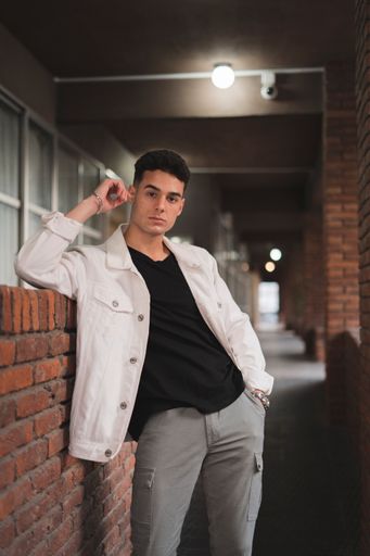 A vertical shot of a young stylish caucasian man in a white jacket leaning on a brick wall