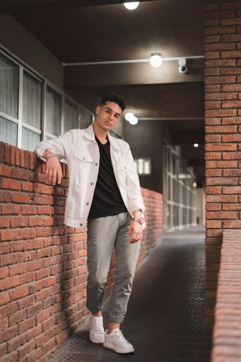A vertical shot of a young stylish caucasian man in a white jacket leaning on a brick wall