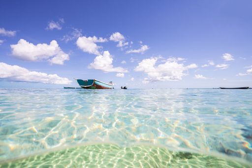 A serene seascape with a traditional boat floating on clear turquoise water under a bright blue sky with fluffy clouds.