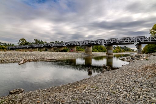A long steel bridge spans over a calm river with a rocky shoreline and a cloudy sky, capturing the serene landscape.