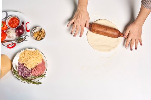 An overhead shot of pizza ingredients and female hands rolling dough on isolated white background