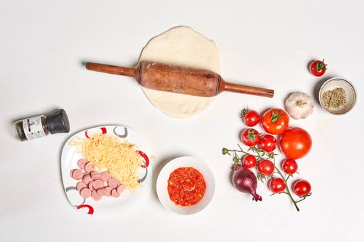 A top view of various ingredients for pizza and a dough roller on a white surface