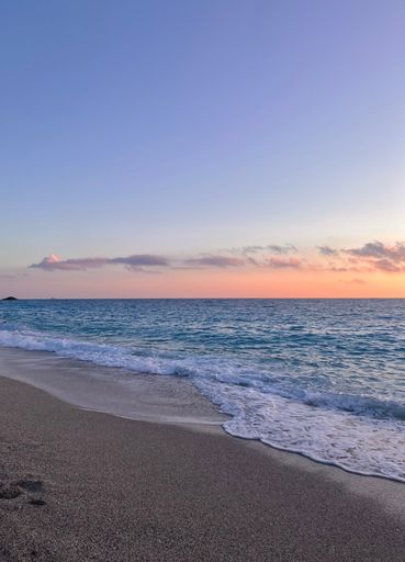 A tranquil beach scene in Greece features gentle waves lapping a sandy shore at sunset. The horizon blends a soft orange glow with a clear blue sky.