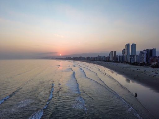 Aerial view of a coastal cityscape at sunset with high-rise buildings and gentle waves on the beach.