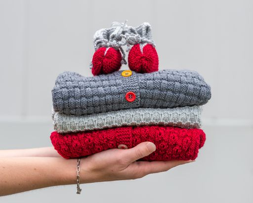 Female hand holding stacked folded baby clothes. Cozy knitted grey and red sweaters with socks on top. Handmade garment to small children.