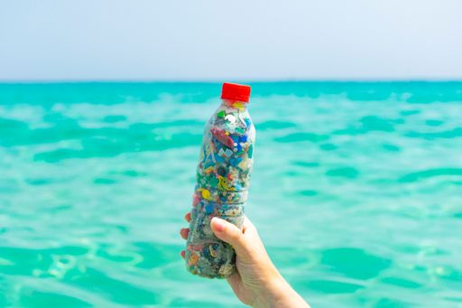 A hand holding a bottle filled with microplastics collected from the sea on the sea background / Concept of saving the environment.