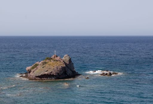 A serene view of a small rocky island with a cross in the middle of the ocean under a clear sky.