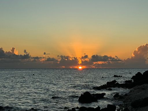 A serene sunset over a rocky shoreline with vibrant orange hues and scattered clouds on the horizon.