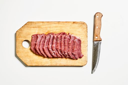 An overhead shot of sliced raw meat on wooden board with knife isolated on white background