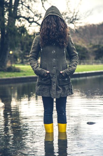 A vertical shot of a female covering her face with hair wearing a raincoat with a hood with her feet in a pond