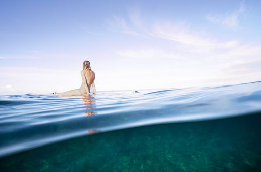A young woman in a swimsuit sits on a surfboard in clear ocean water under a bright sky, capturing a serene moment at sea.