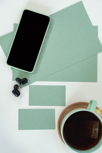 A vertical shot of a smartphone, business card, tea cup, watches, earphones, and paper sheets on a green modern office desk
