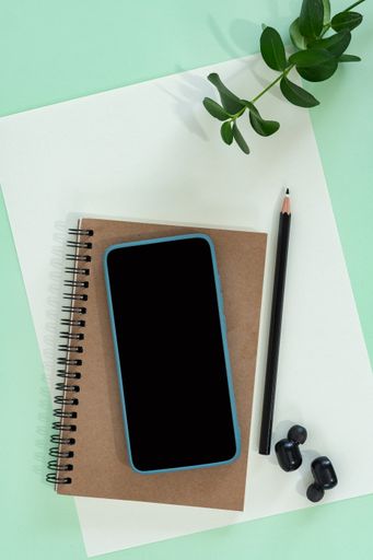 A top view shot of a green surface with an envelope, a notebook, a phone, earphones, and a pencil