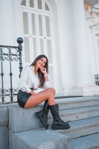 A happy young Caucasian brunette girl in a black leather skirt and a white shirt sitting on the stairs