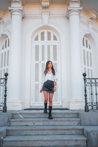 A vertical shot of a brunette female in a black skirt and a white shirt, standing on the stairs in front of the white building