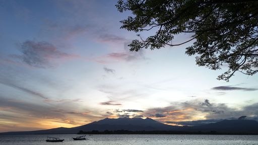 Serene sunset over a mountain landscape with silhouetted trees and boats on the water, creating a peaceful natural scene.