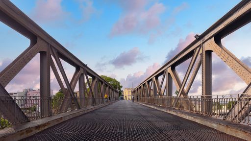 A metal bridge with intricate design under a vibrant sky, featuring two people walking, showcasing urban architecture.