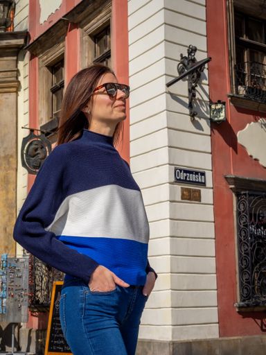 A woman in sunglasses and a striped sweater stands confidently on a sunny street corner in Wroclaw, Poland.