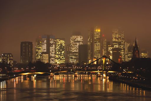 Night view of Frankfurt's skyline with illuminated skyscrapers and reflections on the Main River, showcasing urban architecture.