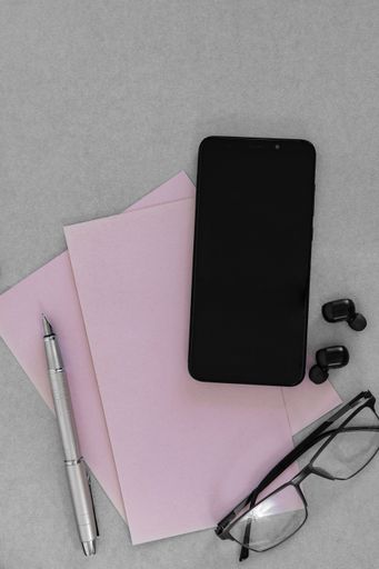 A top view shot of a grey surface with pink papers, a phone, a pen, glasses, and earphones