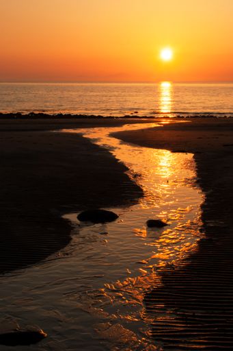 Stream running across Llandanwg beach, Wales at sunset with sun setting over the sea in an orange sky.