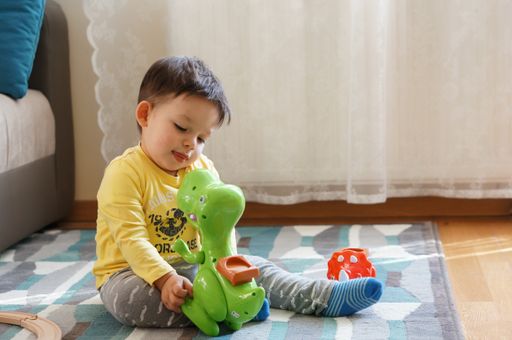A Caucasian little boy sitting on a carpet and playing with a green plastic dinosaur