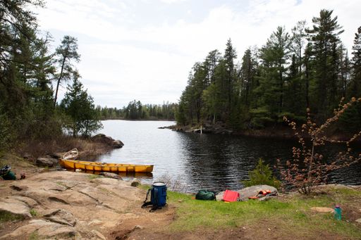 A serene lakeside campsite with a yellow canoe, surrounded by lush green trees and a calm lake under a cloudy sky.