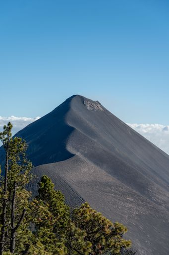 A striking view of a volcanic mountain peak under a clear blue sky with lush green trees in the foreground.