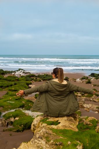 A back view of a woman in a winter coat sitting on the rock with open arms and admiring the ocean
