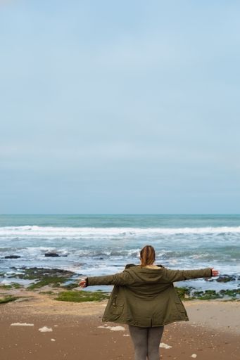 A back view of a woman in a winter coat with open arms admiring the beautiful ocean