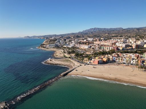 Aerial view of Villajoyosa beach with colorful buildings, clear sea, and distant mountains under a bright blue sky in Spain.