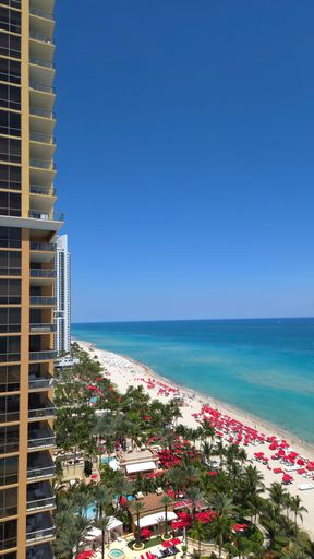 Aerial view of a vibrant beach with red umbrellas and clear blue sea in Miami, Florida, next to a tall building under a cloudless sky.