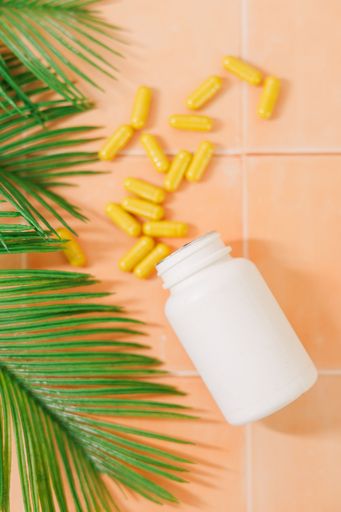 Yellow capsules spill from a white bottle on a peach tile background with green palm leaves.