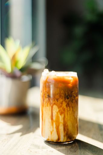 A glass of iced coffee with milk swirls sits on a sunlit wooden table, with a blurred plant in the background.